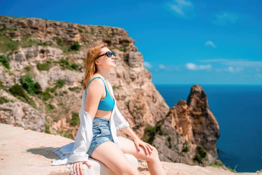 A woman is sitting on a rock overlooking the ocean. She is wearing a blue tank top and white shorts