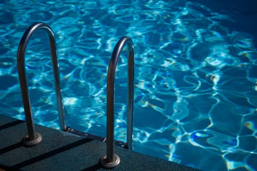 A metal ladder is in the water. The water is clear and calm. The ladder is in the middle of the pool
