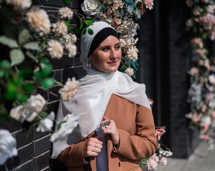 Portrait of a young caucasian woman dressed in a hijab near a wall with flowers