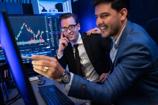 Two stock exchange traders discussing on dynamic successful investment graph, pointing trending of high profit in currency stock in financial data on monitor in neon light at workplace. Sellable.