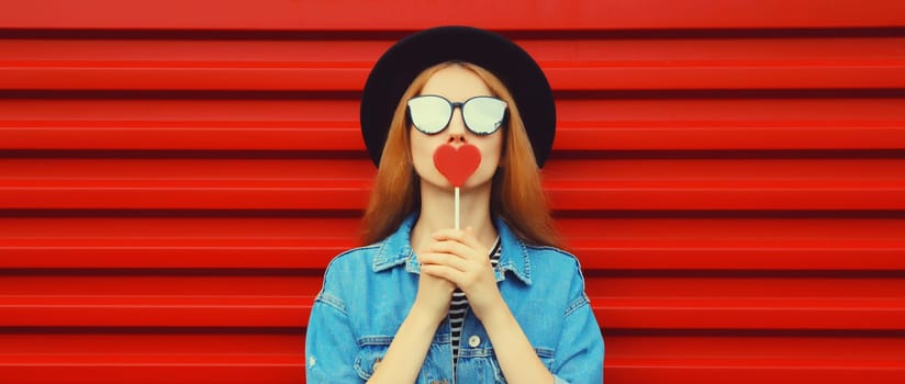 Portrait of beautiful young woman with lollipop blowing kiss wearing sunglasses, black round hat on red background