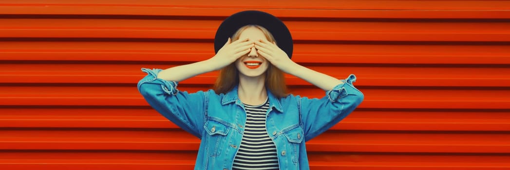 Portrait of happy surprised smiling young woman covering her eyes wearing black round hat, denim jacket on red background