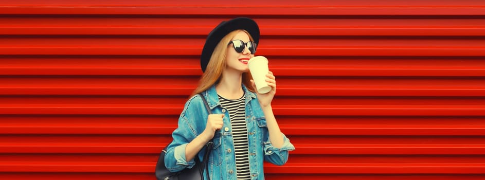 Beautiful stylish happy young woman with cup of coffee wearing black round hat, blue jacket and backpack on red background