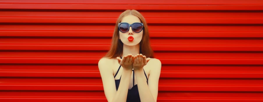 Portrait of stylish caucasian young woman model blowing a kiss wearing in black outfit, sunglasses on red background