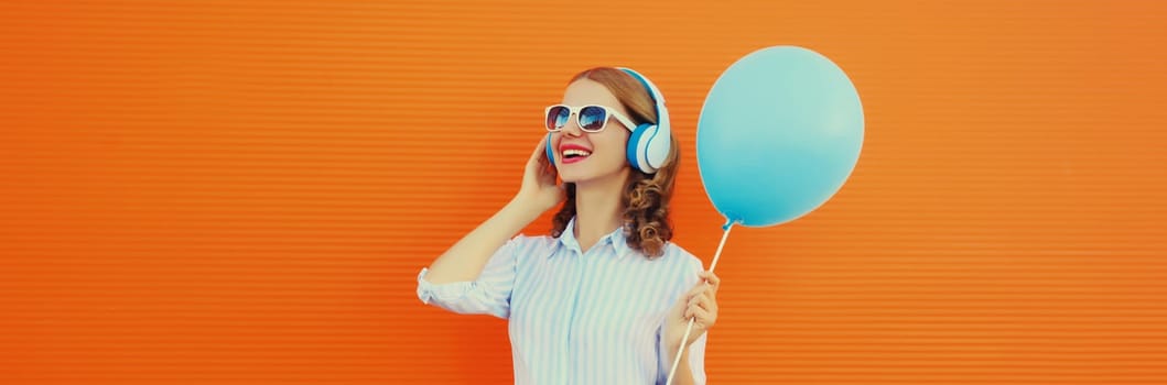 Portrait of happy cheerful young woman in wireless headphones listening to music with blue balloon on orange background