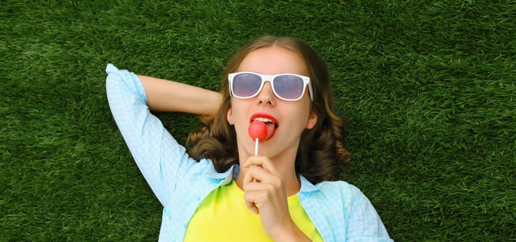 Portrait of happy teenager girl resting with lollipop lying on green grass in summer park, top view