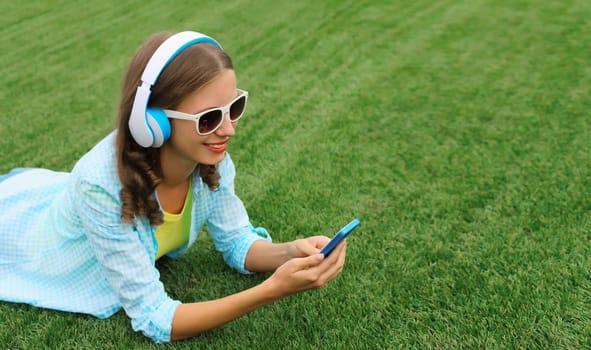 Happy young woman holding phone listening to music in headphones while lying on grass in summer park