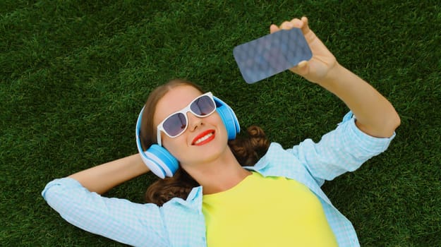 Happy young woman taking selfie with smartphone listening to music in headphones while lying on grass in summer park