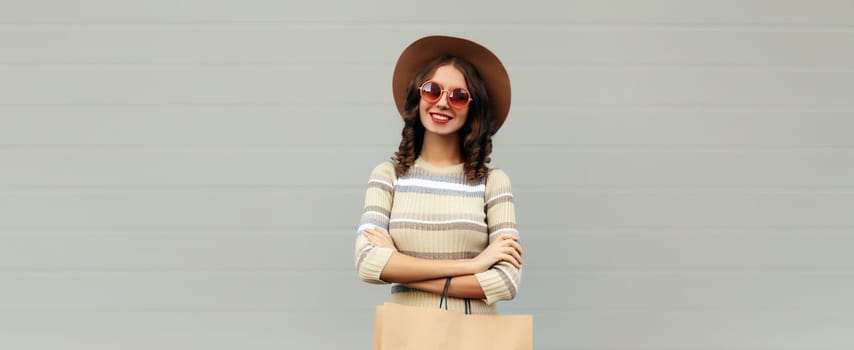 Stylish beautiful happy smiling young woman posing with shopping bags in round hat, dress on city street, gray wall background