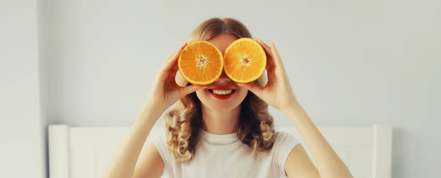 Summer, nutrition, diet and vegetarian concept. Happy healthy cheerful young woman covering her eyes with slices of orange fruits and looking for something in white room at home