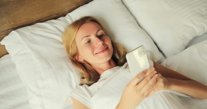 Healthy sleep, happy relaxed young woman with mobile phone lying in bed on white soft comfortable pillow at home