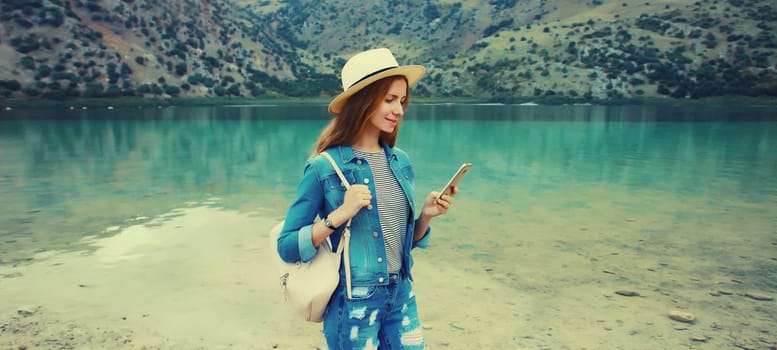 Summer vacation, young woman tourist using mobile phone wearing straw hat with backpack on lake and mountains background. Greece, island Crete
