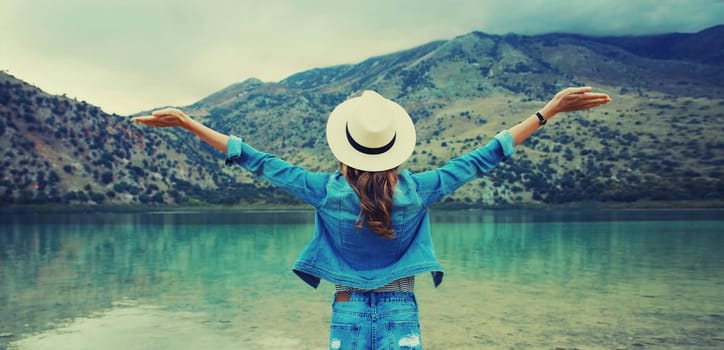 Summer vacation and tourism, rear view of happy woman tourist in straw hat raising her hands up on lake and mountains background. Greece, island Crete.