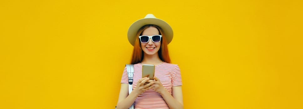 Summer portrait of happy traveler young woman 20s with mobile phone looking at device in casual straw hat with backpack on bright yellow studio background