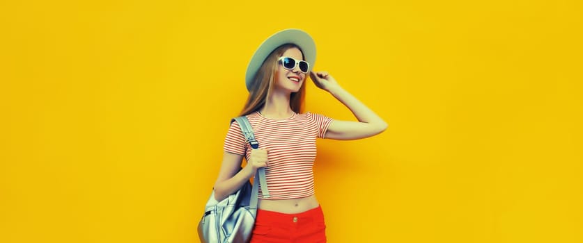 Beautiful young woman posing in summer straw hat, red shorts with backpack looking away on vivid yellow studio background