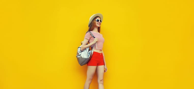 Beautiful young woman full length standing in summer straw hat, red shorts with backpack on vivid yellow background