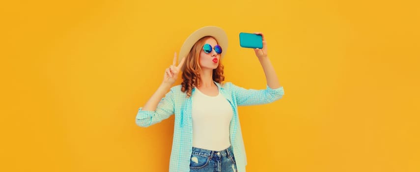 Summer cheerful stylish young woman taking selfie with smartphone blowing a kiss wearing straw hat on yellow background