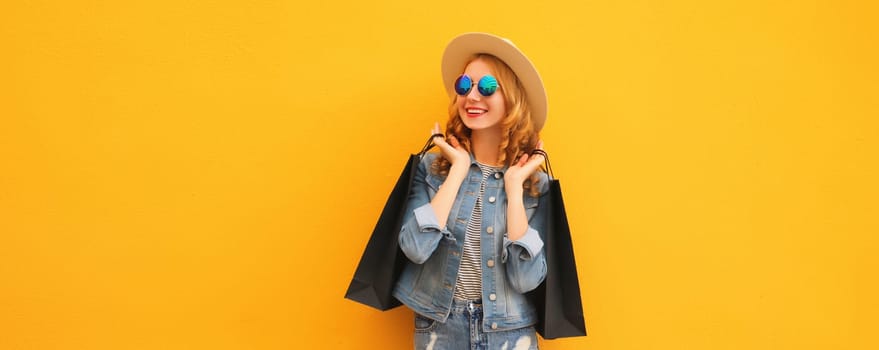 Shopping day! Stylish beautiful happy smiling young woman posing with black shopping bags wearing denim clothing, summer straw hat on yellow studio background