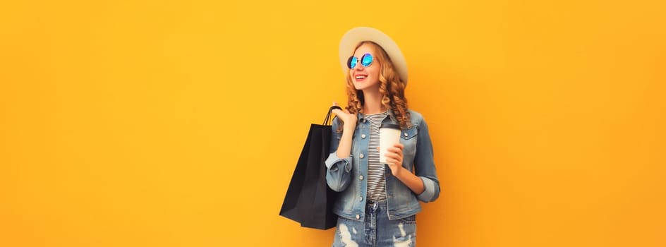 Shopping day! Stylish beautiful happy smiling young woman posing with black shopping bags wearing denim clothing, summer straw hat on yellow studio background