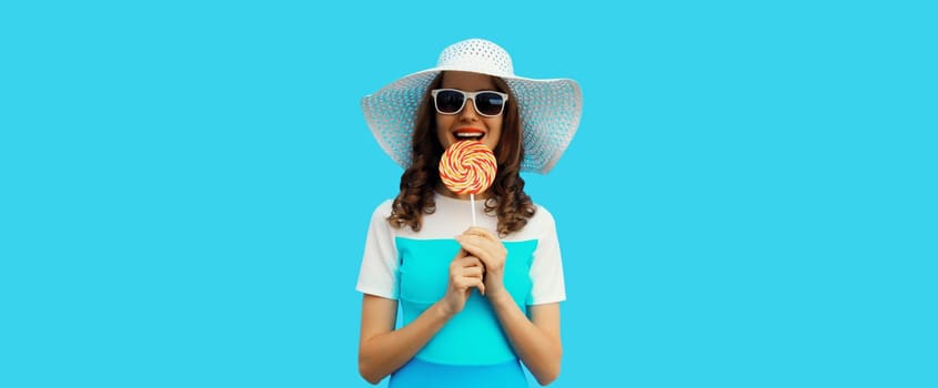 Portrait of happy cheerful smiling young woman holding colorful lollipop wearing white summer hat, sunglasses on blue background