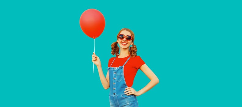 Happy cheerful young woman having fun with balloon wearing red heart shaped sunglasses, jumpsuit on blue background