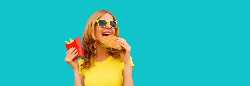 Portrait of happy cheerful young woman eating burger fast food and french fries, fried potatoes isolated on blue studio background