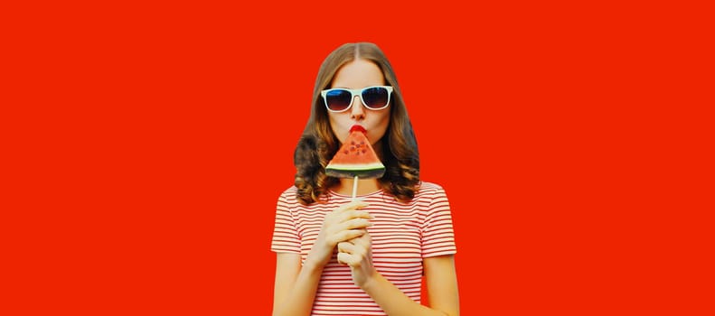 Summer portrait of stylish young woman with sweet juicy lollipop or ice cream shaped slice of watermelon wearing glasses on red studio background