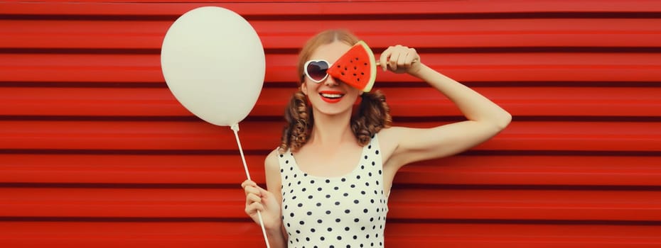 Happy cheerful young woman with balloon and fresh fruit lollipop or ice cream shaped slice of watermelon wearing white heart shaped sunglasses on red background