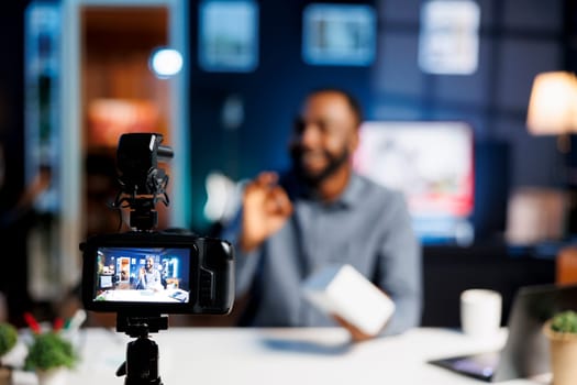 Focus on professional video production camera used for capturing footage of content creator in blurry background doing products unboxing for online audience, showcasing different tech electronics