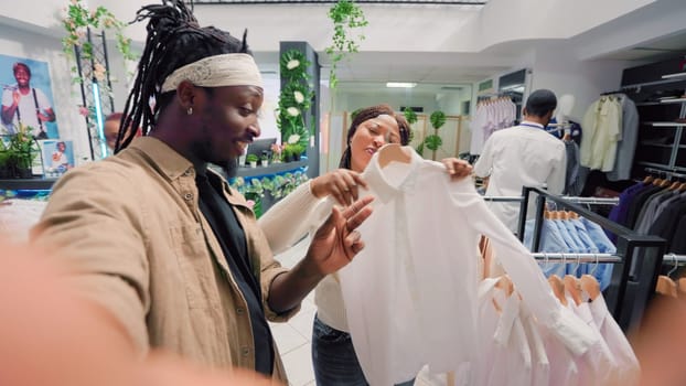 Man filming POV vlog with his partner while doing shopping in elegant clothing store, giving fans info. Fashion influencer promoting new collection stock on social media with girlfriend