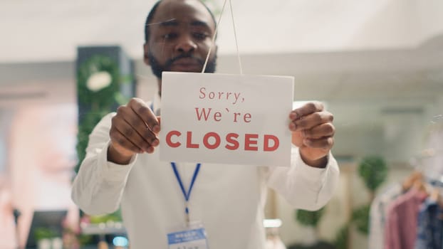 African american man working in trendy clothing store closing it at night. Tired BIPOC retail clerk turning opened message sign on fashion boutique door in mall at shift end