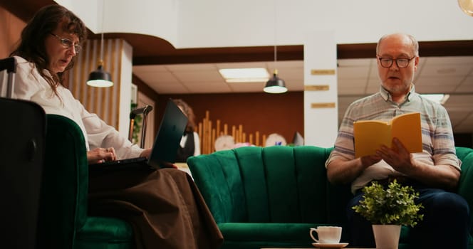 Senior couple passing time at hotel, wife creating trip itinerary on laptop while husband reading book in lounge area. Retired people travelling on international vacation, waiting for check in.