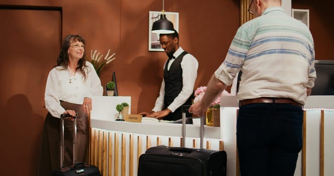 Retired couple arriving at hotel reception to see room reservation, travelling on international vacation with all inclusive services. Husband and wife talking to staff about senior travel offers.