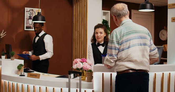 Elderly person arriving at hotel front desk with trolley bags, asking for assistance to check in. Receptionist ensuring pleasant stay for old man at all inclusive holiday retreat, tourism industry.