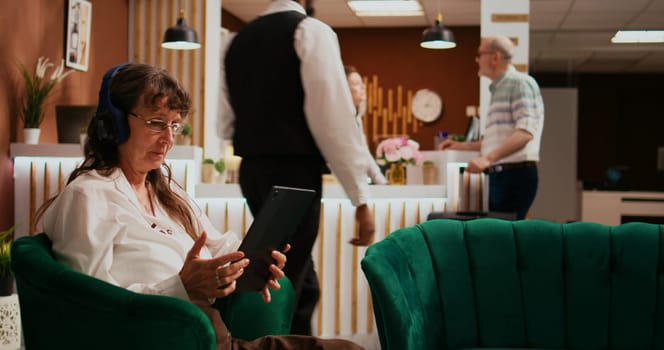 Retired traveller using tablet in lobby, watches film on streaming services while she waits to see accommodation. Elderly woman enjoying modern technology with gadget and headphones.