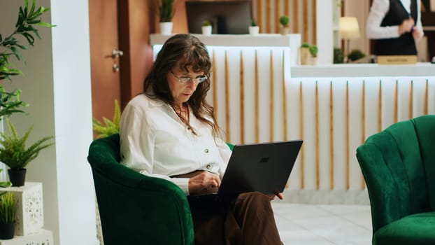 Senior tourist using laptop in lounge area, navigating internet to look at visiting places on website. Hotel guest arrives at holiday retreat and waiting for room reservation. Handheld shot.