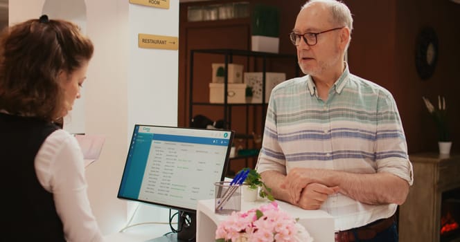 Receptionist looks at passport info to check id of senior adult arriving at luxurious hotel, retirement age holiday. Woman staff helping tourist with check in process, welcoming man at resort.