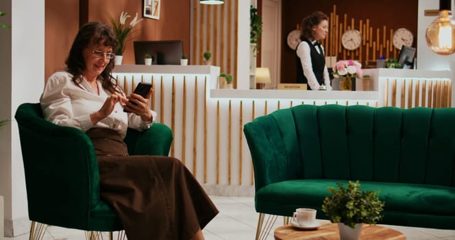 Old woman reads text messages after international flight, waiting for check in procedure at five star resort. Senior tourist browsing smartphone app in lounge area, relaxing in luxurious lobby.