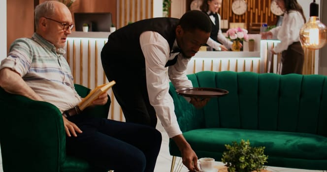 Client paying cash for coffee cup in lounge area at five star hotel, giving money to african american bellboy. Elderly adult reading literature book in lobby and receiving drink from bar.