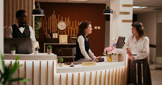 Elderly person arriving in hotel lobby, asking concierge about senior travel offers for wellness and leisure activities. Employee welcoming tourist at reception, ensuring relaxing stay.