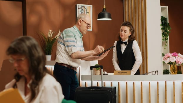 Elderly man signs digital forms for room check in at hotel front desk, talking to receptionist about leisure activities to ensure pleasant trip. Employee greeting visitor at reception. Handheld shot.