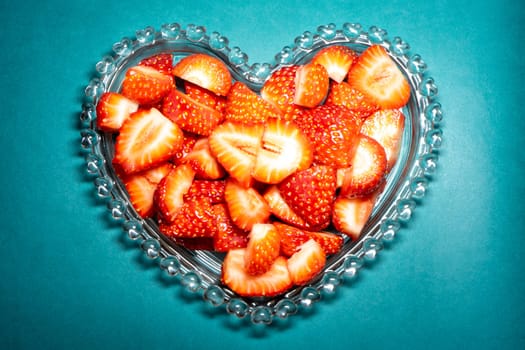 Peaces of strawberries in a heart shaped bowl on a blue background.