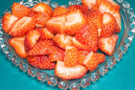 Peaces of strawberries in a heart shaped bowl on a blue background.