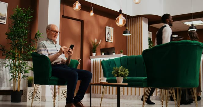 Retired traveller browses smartphone app in lounge area at five star holiday retreat, using online network to navigate on webpage. Senior client at hotel relaxing in lobby.