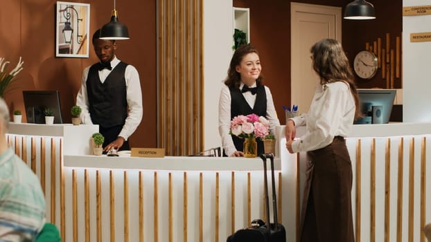 Female client checking in at luxury hotel lobby with assistance from friendly concierge staff. Receptionist greeting senior woman at front desk and helping with registration. Handheld shot.