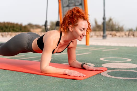 Happy fit strong woman with red hair doing plank exercise on mat. Side view of female athlete in sportswear doing fitness exercise outdoors on sunny summer day.