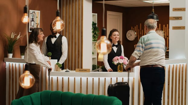 Senior couple arrive in hotel lobby, talking to front desk employee before check in process. Tourists travelling on vacation for leisure activities, hotel concierge services. Tripod shot.