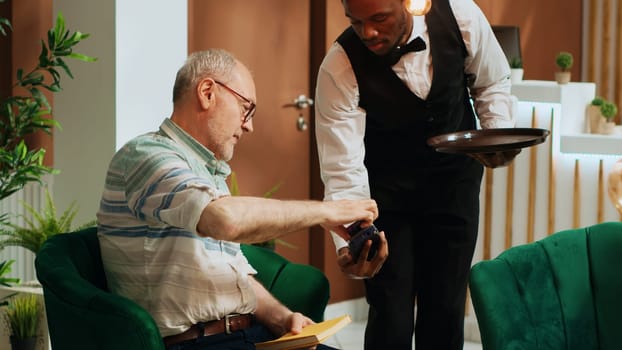 Elderly person paying for coffee cup via credit card in hotel lobby, ordering refreshment from bar and making pos transaction. Bellhop serving guest with beverage while he is waiting.