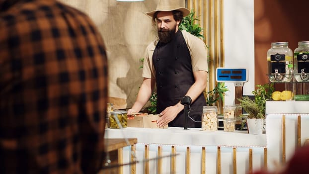 Woman at zero waste supermarket checkout counter taking groceries out of paper bag for vendor to weight on electronic scale. Storekeeper measuring customer groceries kilograms before payment