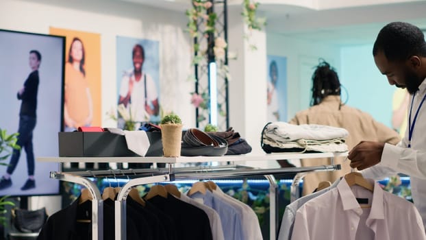 African american employee working in SH clothing shop, placing simple stylish shirts on racks. Retail assistant formal garments merchandise on hangers in discount fashion store